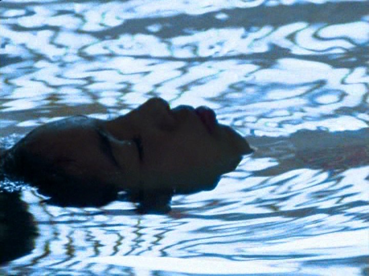 A tightly cropped image shows a child’s head emerging from a pool. The person is lying on their back, with their ears still underwater. A pattern reflected on the water’s surface appears to come from the roof structure above.