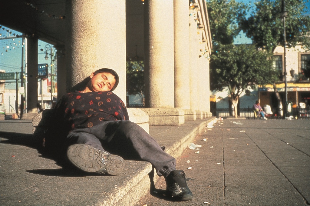 Eine Person liegt in einer urbanen Szene gegen die Säule eines Monuments gelehnt mit geschlossenen Augen auf dem Boden.