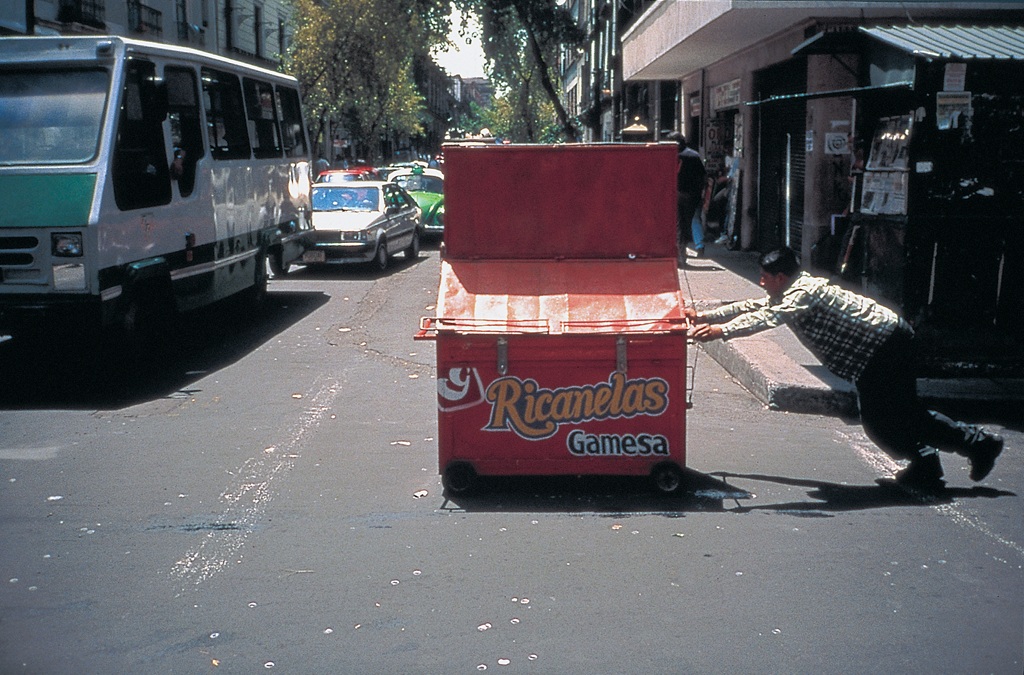 In einer verkehrsreichen Straßenszene scheint ein Mann einen roten Müllcontainer von Hand quer über die Straße zu schieben.