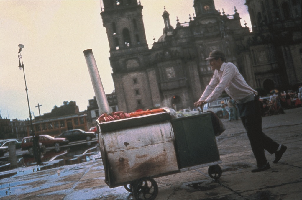 Eine Person schiebt einen Imbiss-Wagen über einen Platz mit einem historischen Gebäude im Hintergrund. 