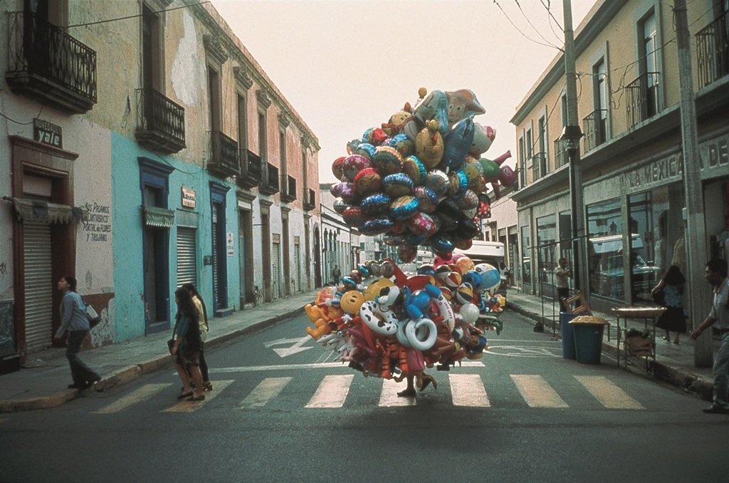 Eine Person trägt große Bündel Heliumluftballons über einen Zebrastreifen und verschwindet dabei bis auf die Füße hinter der Fülle der Ballons.