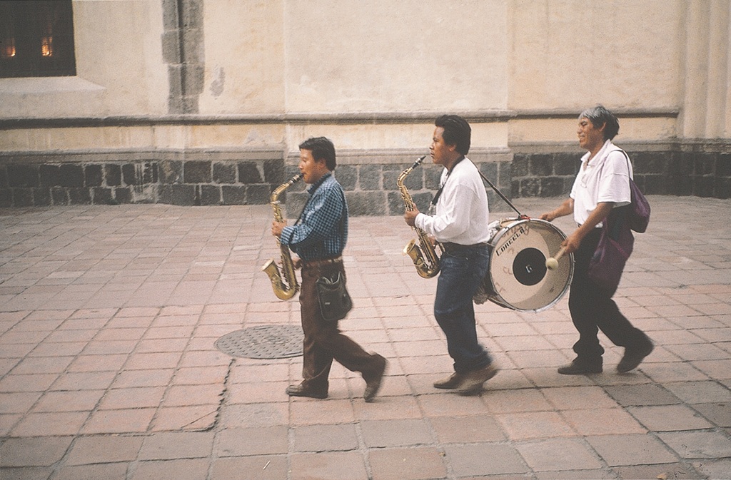 Zwei Saxophonspieler und ein Trommler laufen musizierend die Straße hinunter, im Hintergrund befindet sich ein Gebäude.