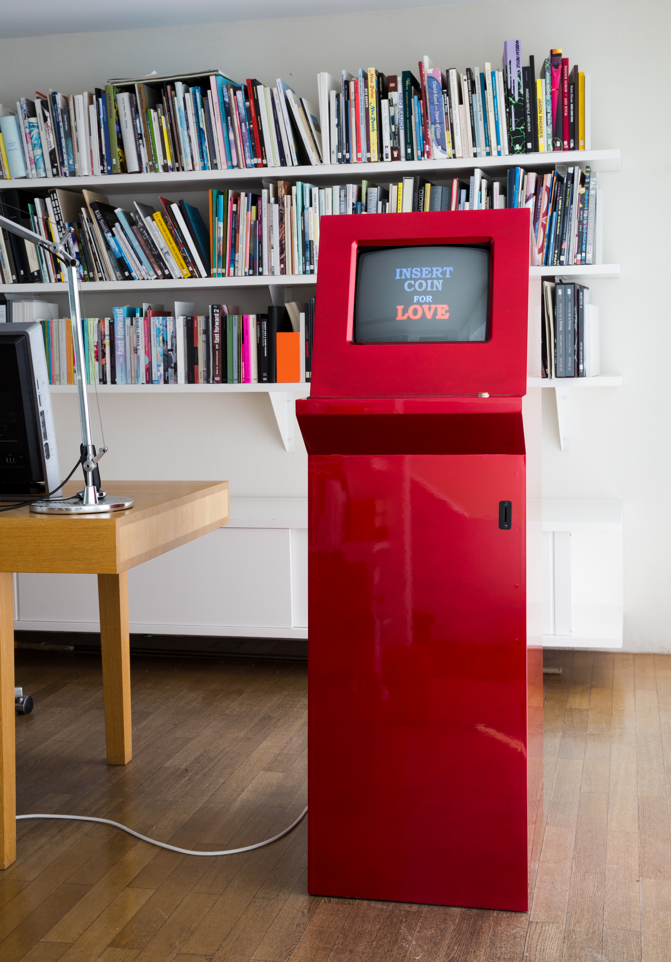 A red coin-operated machine with a monitor stands in front of a bookshelf