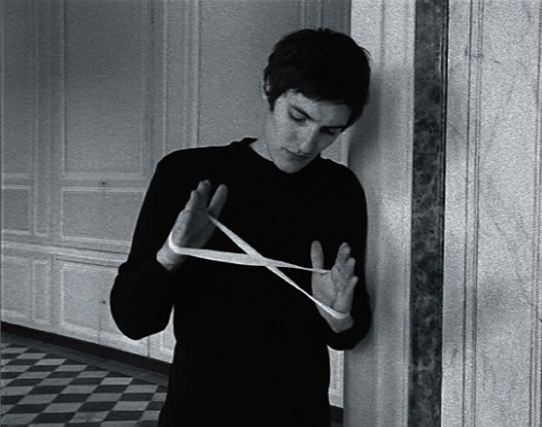 Schwarzweiß-Aufnahme: ein junger Mann hält ein weißes Gummi- oder Stoffband überkreuzt zwischen den Händen, so dass es eine 8-Form annimmt.