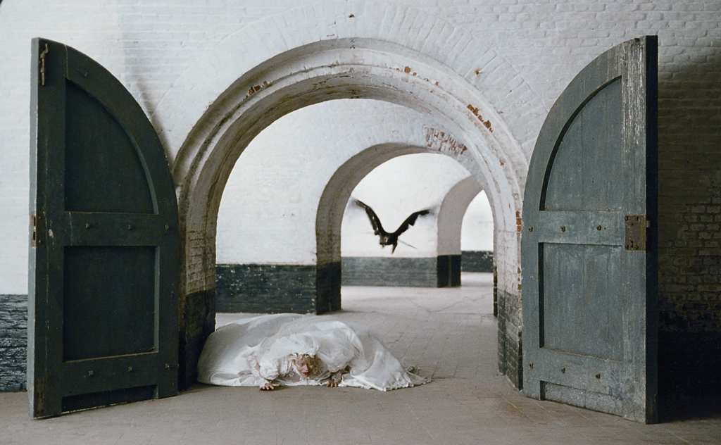 A blonde woman in a wedding dress lies on the floor under a barrel vault made of white bricks. A bird of prey (raven or owl) flies over her.