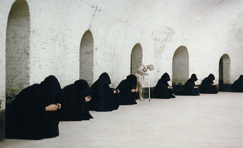 People with black hooded capes sit in a semicircle in front of white brick walls. A white figure with a walking stick strides through them. 