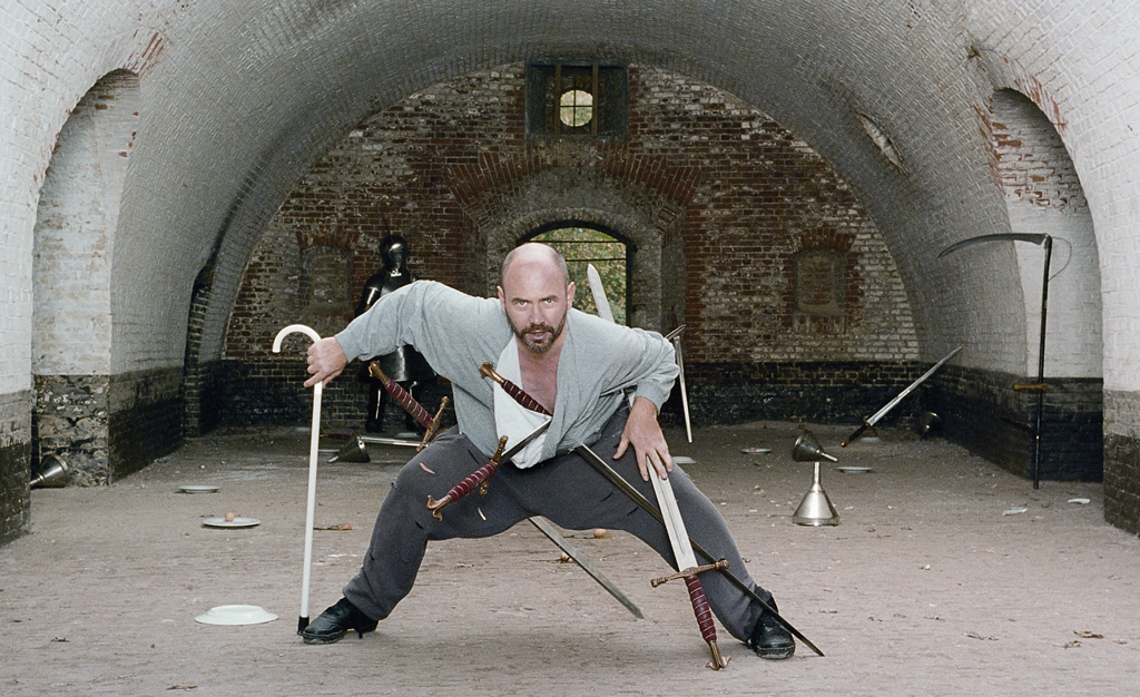 Under a barrel vault made of white bricks, a man stands in a kind of attack stance with his legs apart and knees bent. Several swords are attached to his clothing and he is leaning on a walking stick with his right hand. Armor and a scythe can be seen in the background, as well as other metal objects. 