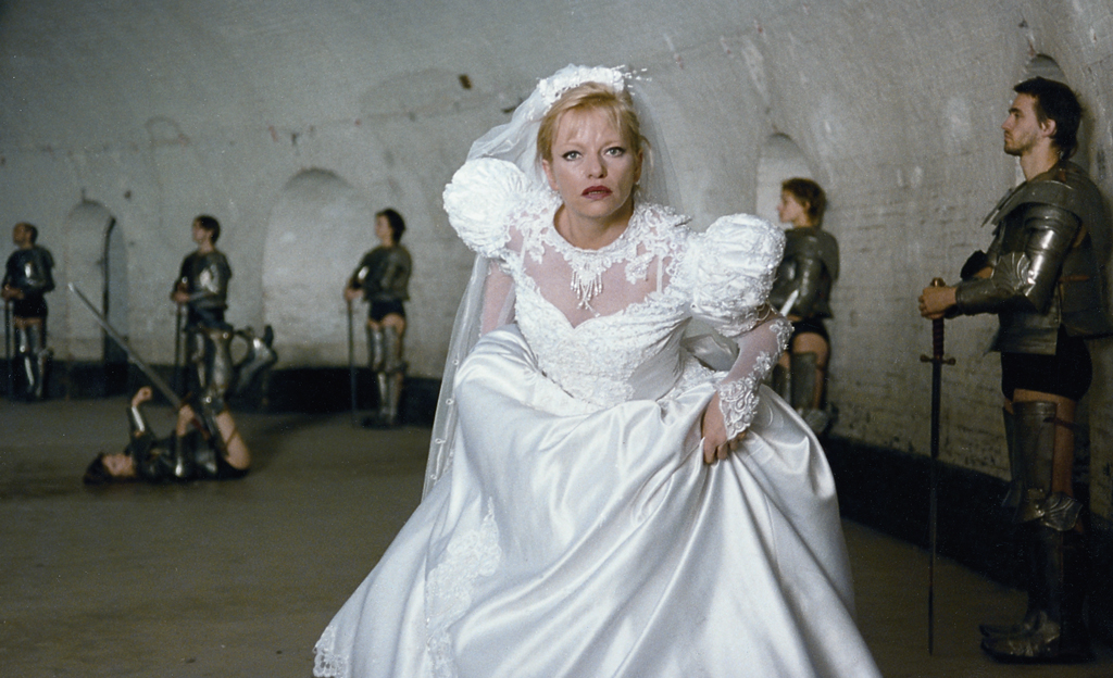 A blonde woman in a wedding dress appears to be on the run. Behind her are people in knight's armor with swords in front of a white brick wall. One of them is lying on the ground, her sword raised in the air.