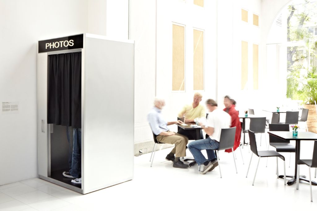 A silver aluminium photo booth stands in a brightly lit courtyard. Behind the closed black curtain, only the legs of two people in jeans are visible, standing so close together that it gives the impression the pair are kissing. Next to the photo booth is a table where a group of people is seated.