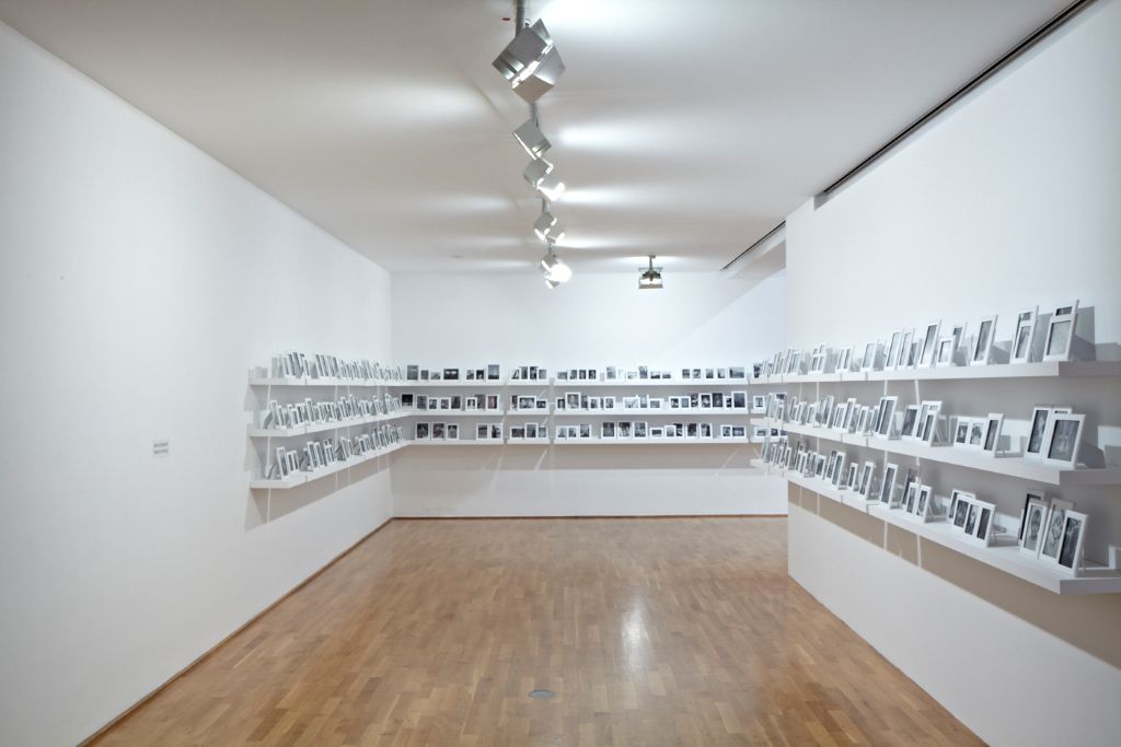 Three rows of shelves wind around the corner of a corridor. On the shelves are photographs in white frames. The photographs mainly depict young people, mostly male couples.