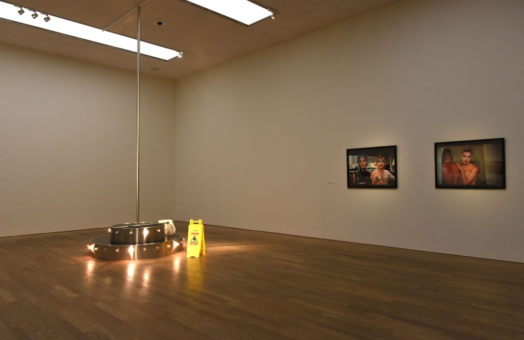 In an exhibition room, where two colour photographs hang on the walls, stands a pole-dancing pole. Two circular platforms outlined with light bulbs lead up to the pole. Beside them are a mop bucket and a yellow warning sign indicating a wet floor.