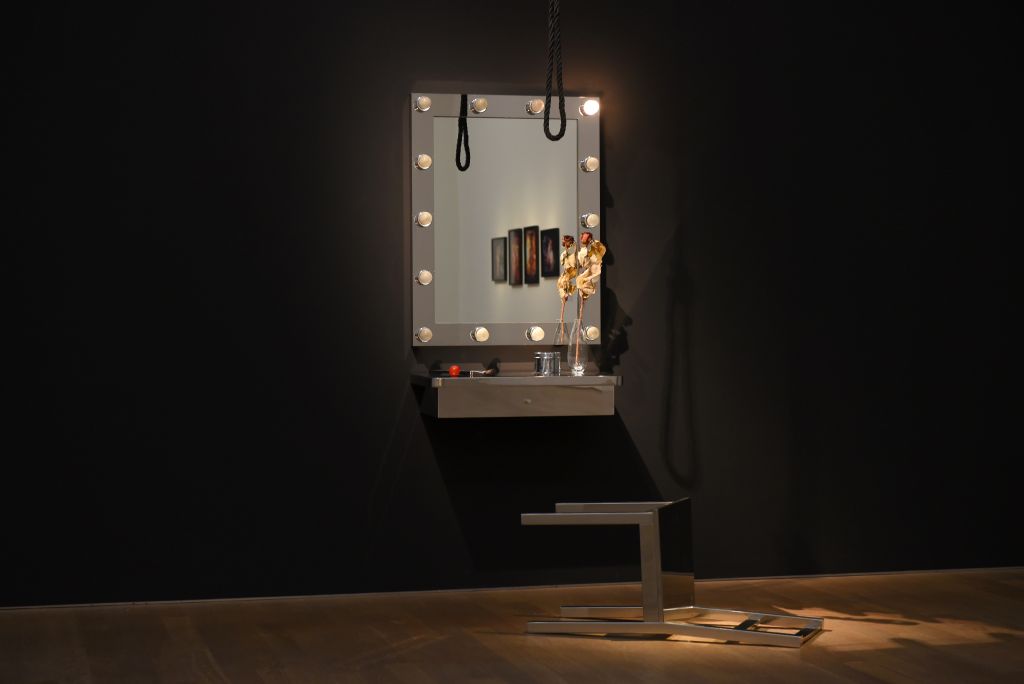 An einer dunkel gestrichenen Wand hängt ein Spiegel, der von Glühbirnen umrandet ist. Davor liegt ein umgekippter Stuhl. Von der Decke hängt ein Seil in Schlaufenform.