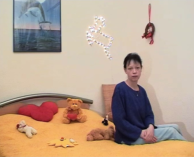 Auf einem gelb bezogenem Bett sitzt eine Frau in blauem Pullover. Sie hat ihre Hände in den Schoß gelegt. Auf dem Bett liegen verstreut Kuscheltiere und hinter dem Bett an der Wand hängt ein gerahmtes Delfinposter.