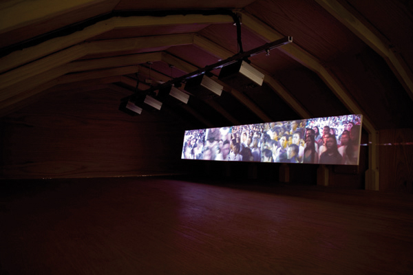 Under a wooden vault stands a long screen on which 5 projectors project images of a dense group of people. 