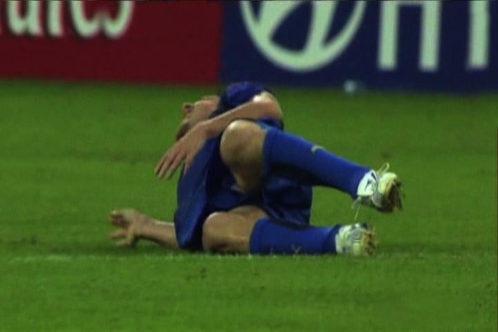 Ein Fußballspieler in rotem Trikot fällt wie von unsichtbarer Hand auf ein sattgrünes Fußballfeld. Im Hintergrund sind Werbebanner zu sehen.