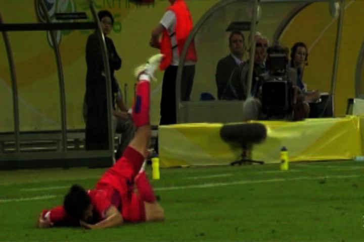 Ein Fußballspieler in rotem Trikot fällt wie von unsichtbarer Hand auf ein sattgrünes Fußballfeld. Im Hintergrund sind Kameras zu sehen.