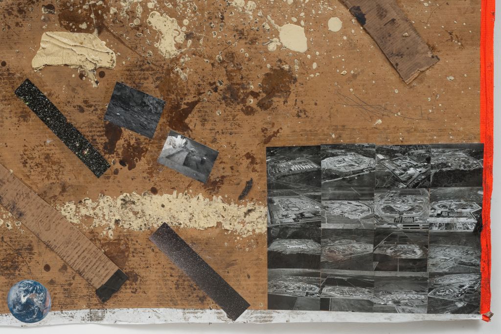 A detail of the image shows a piece of cardboard. The surface of the cardboard, which is partially marked by pale yellow paint splatters and dirt, is covered with small black-and-white photographs. Some depict landscapes; in the lower right corner, several photographs of fortified structures are arranged into a rectangular formation.