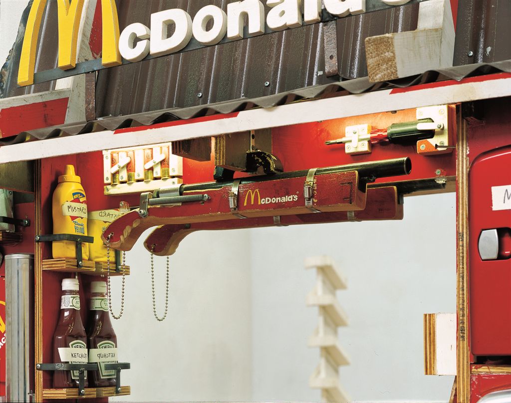 Bildausschnitt einer größeren im McDonalds-Stil gehaltenen Objektstruktur. Am oberen Bildrand ist ein mit Dachpappe gedecktes Dach zu sehen, auf dem der McDonalds-Schriftzug mit dem charakteristischen geschwungenen gelben „M“ angebracht ist. Unter dem Dach, vor einem Spiegel, der anstelle eines Drive-Through-Fensters montiert ist, hängt ein rot gestrichenes Holzgewehr mit McDonalds-Schriftzug. Dahinter sind Patronen in einer hölzernen Halterung befestigt. Links daneben stehen in ebenfalls aus Holz gefertigten Vorrichtungen eine Ketchup- und eine Senfflasche.