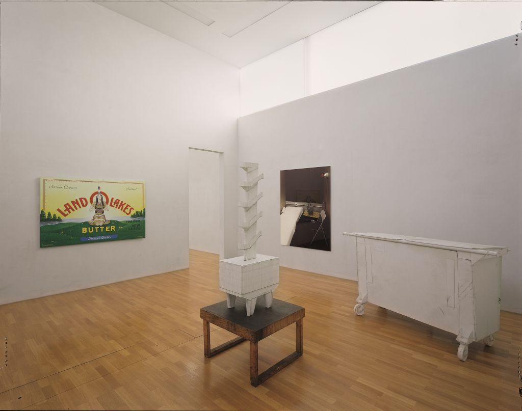 In einem hellen Raum mit einem Lichtbandfenster und Parkettboden sind vier Kunstobjekte platziert. In der Raummitte stehen zwei Skulpturen, die in Weiß gehalten sind. Sie sind aus Schaumplatten angefertigt. Die eine Skulptur steht auf einem Sockel und stellt einen aufgebockten Gebäudekorpus dar, aus dem eine viergeschossige Treppe hinausragt. Die zweite größere Skulptur ahmt die Form eines Müllcontainers nach. 