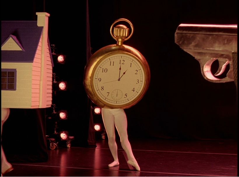 Auf einer Bühne steht eine  altmodische Taschenuhr auf zwei Ballerina-Beinen. Neben ihr stehen, ebenfalls auf Tänzerbeinen, ein Haus und eine Pistole.