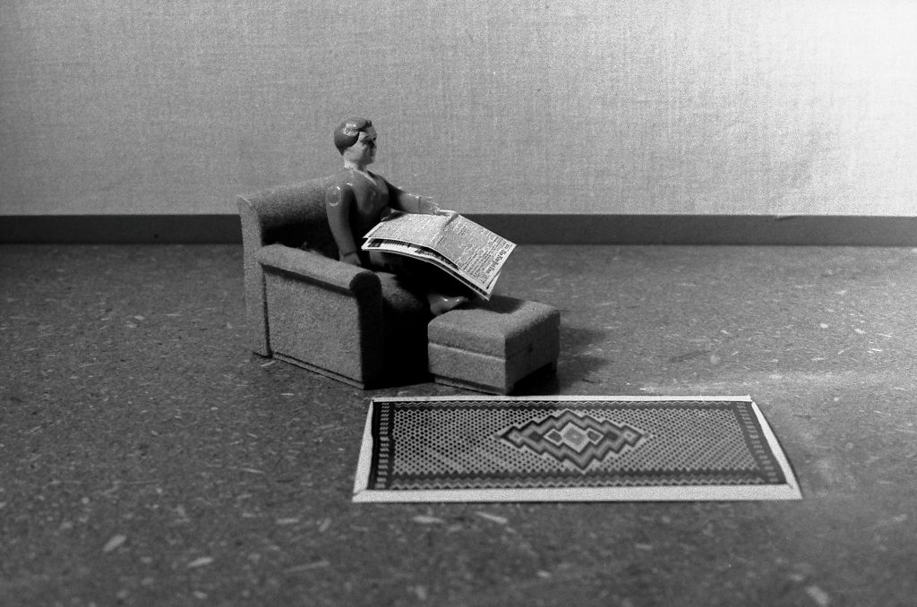 A female doll figure sits in a simple room in an armchair with an ottoman. She holds a miniature newspaper in her hand. In front of the chair lies a patterned woven rug. The photograph is rendered in black and white.
