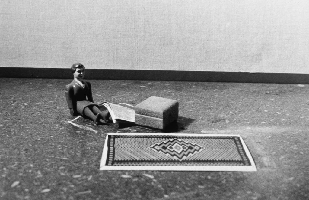 A female doll figure sits on the floor in a simple room, holding a newspaper. An ottoman stands in front of her. At the spot where an armchair could be placed, there is a chalk outline. In front of her lies a patterned woven rug. The photograph is rendered in black and white.