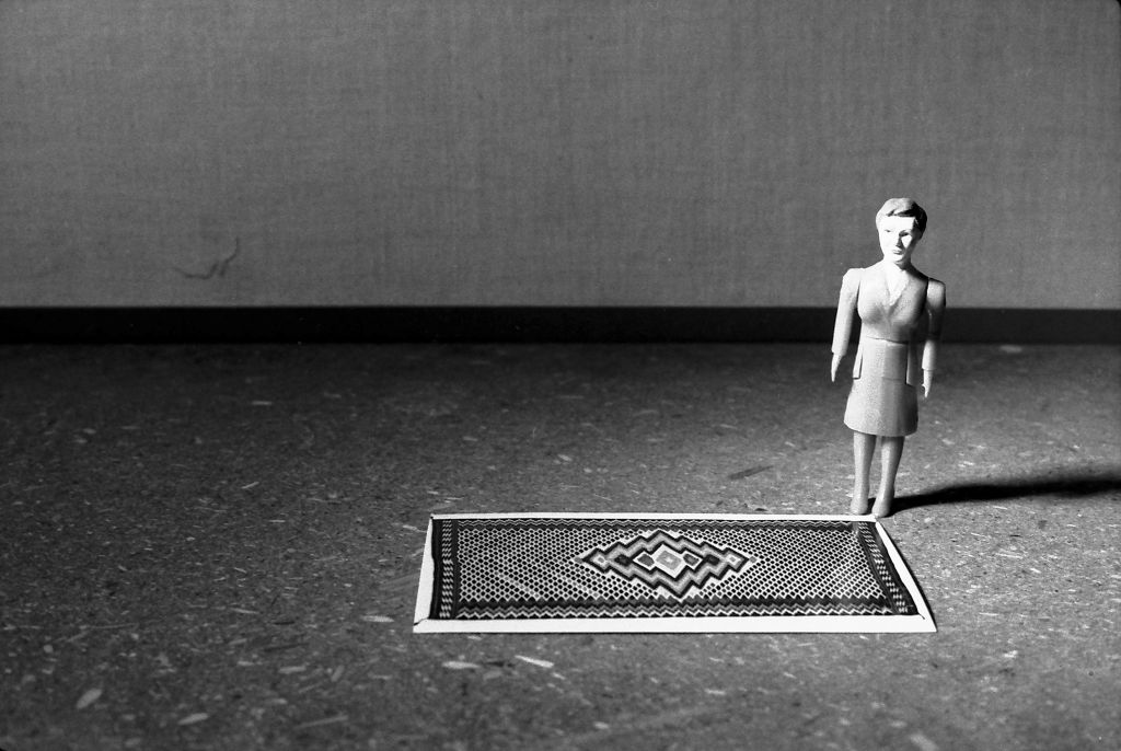 A female doll figure wearing a suit stands in a simple room. In front of her lies a patterned woven rug. She stands at its right edge. The photograph is rendered in black and white.