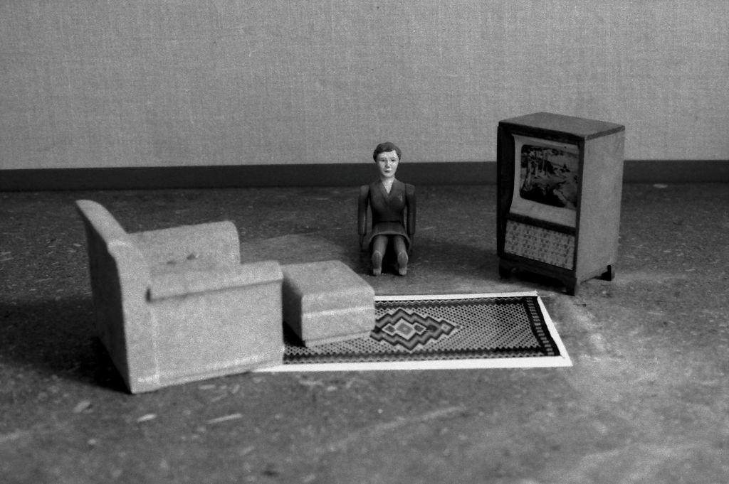 A female doll figure wearing a suit sits on the floor of a living room. She faces the camera directly, with her legs stretched straight out. The room contains an armchair with an ottoman, a patterned woven rug, and a television housed in a wooden cabinet. The photograph is rendered in black and white.
