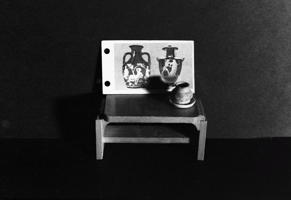 The photograph shows miniature furniture against a dark background. On a small table stand a ceramic vase and an image depicting Greek vases. The photograph is rendered in black and white. 