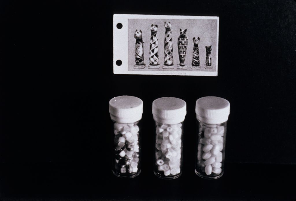 The photograph is rendered in black and white and shows three transparent cylindrical pill containers against a black background. Behind the containers, an image depicting five sarcophagus-like figures on plinths is mounted.