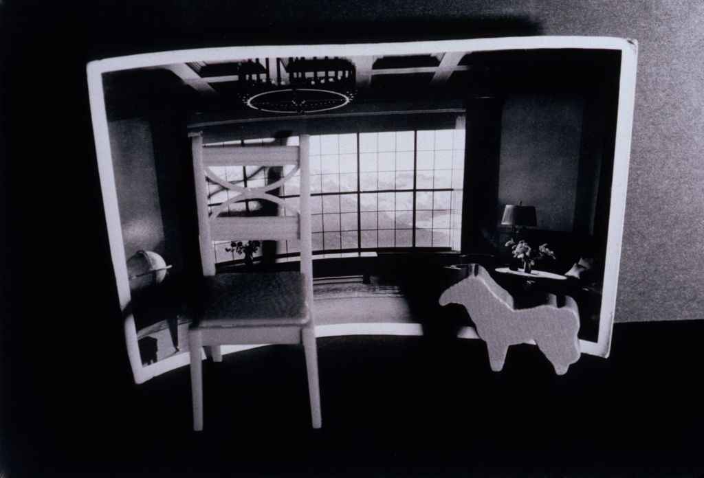 The black-and-white photograph shows a toy chair and a small toy horse in front of another photograph. The latter depicts the view from a spacious living room overlooking an Alpine panorama.