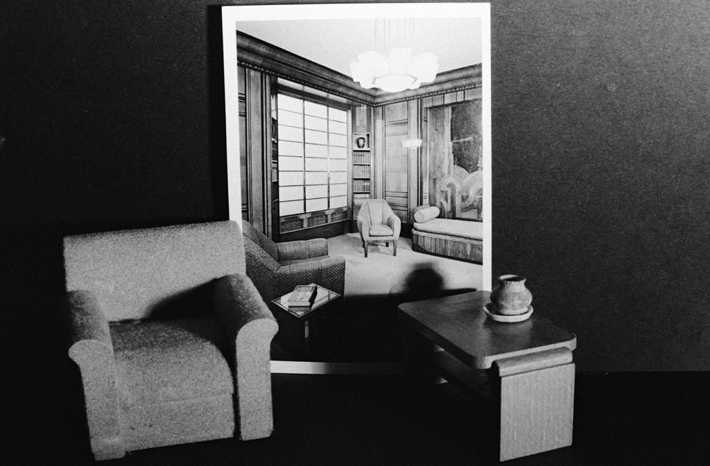 The black-and-white photograph shows a toy armchair next to a toy table on which a small ceramic vase is placed. In the background is a photograph of a bourgeois living room with an armchair, a chair, and a recamier. The walls are panelled in wood.