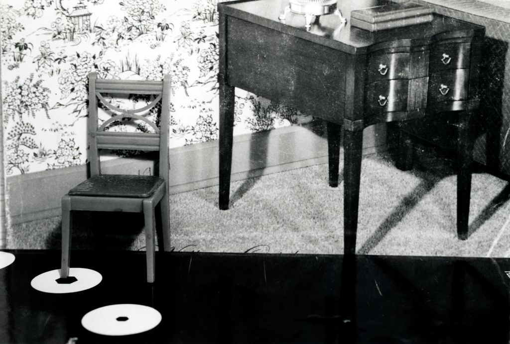 A toy chair stands in front of a photograph of a bourgeois interior featuring Biedermeier furniture, a high-pile carpet, and floral wallpaper. The toy furniture and the furniture in the photograph are approximately similar in scale.