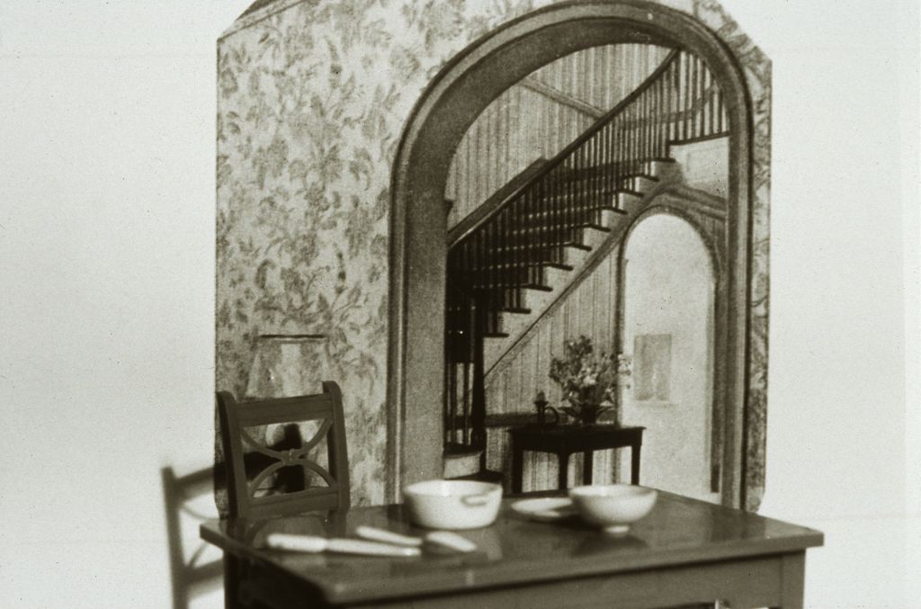 The black-and-white photograph shows a set toy table placed in front of another photograph depicting a spacious staircase. The proportions are aligned in such a way that it appears as though the toy table belongs to the architectural space shown in the background.