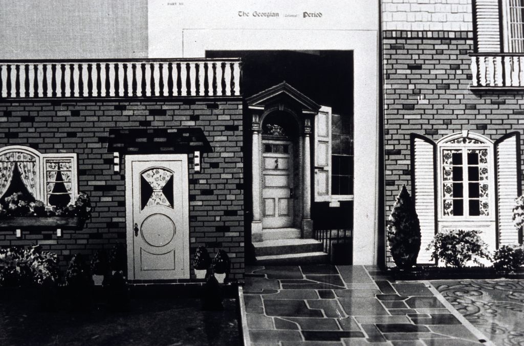 The entrance of a house has been collaged from various printed cardboard boxes and newspaper clippings. The doorway is formed by an excerpt from a textbook titled The Georgian Period. The photograph is rendered in black and white.