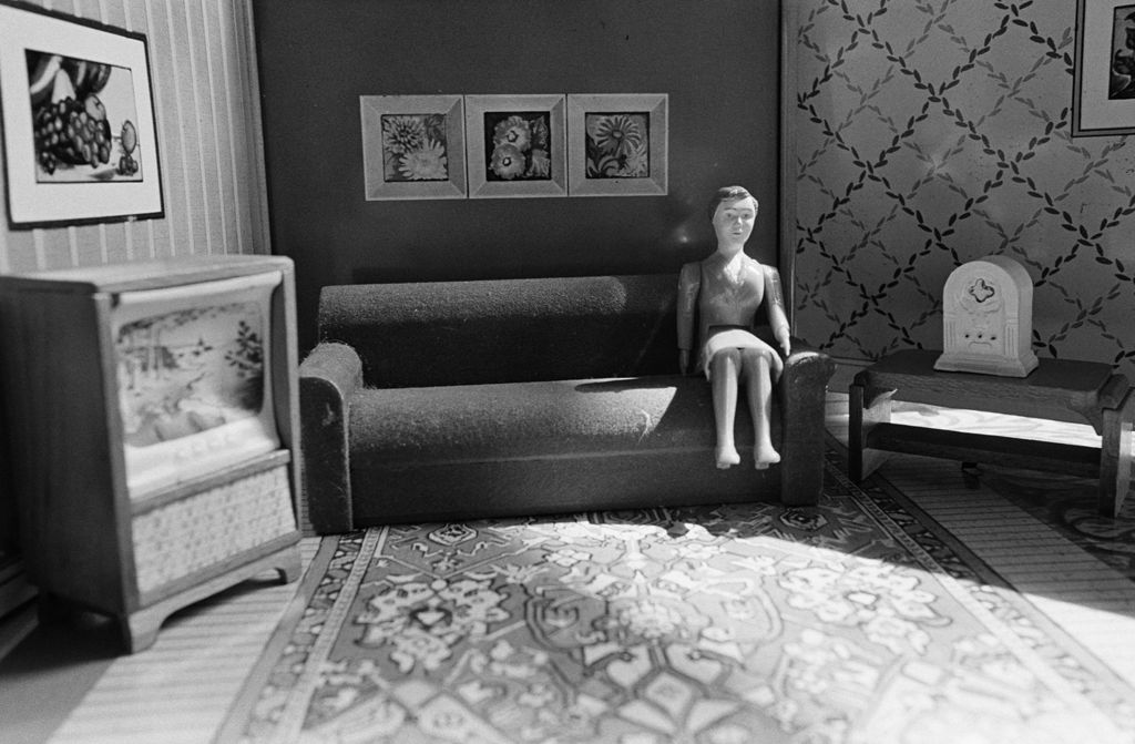 The photograph shows the interior of a dollhouse. The walls are painted in different colours and decorated with floral still lifes. In front of a wall with three pictures stands a sofa on which a doll figure is seated. Next to the sofa are a television in a wooden cabinet and a clock on a wooden table. Beneath the sofa, extending across the entire room, lies a large carpet. The photograph is rendered in black and white.