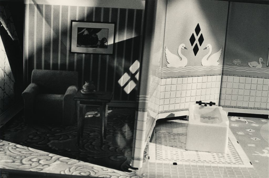 The black-and-white photograph shows a view into a dollhouse. The image is composed in cross-section, allowing two rooms to be seen simultaneously. One half shows a wallpapered living room with an armchair and a small side table. The other half shows a tiled bathroom with a bathtub. The tiles are installed only up to waist height; the upper section is covered with swan-patterned wallpaper.