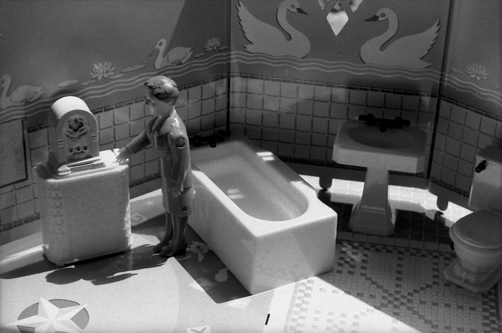 The black-and-white photograph shows the interior of a miniature bathroom. Inside the bathroom, a female doll figure stands in front of a dresser on which an old-fashioned radio is placed. Behind her, additional miniature bathroom fixtures are visible, including a bathtub, a sink, and a toilet.