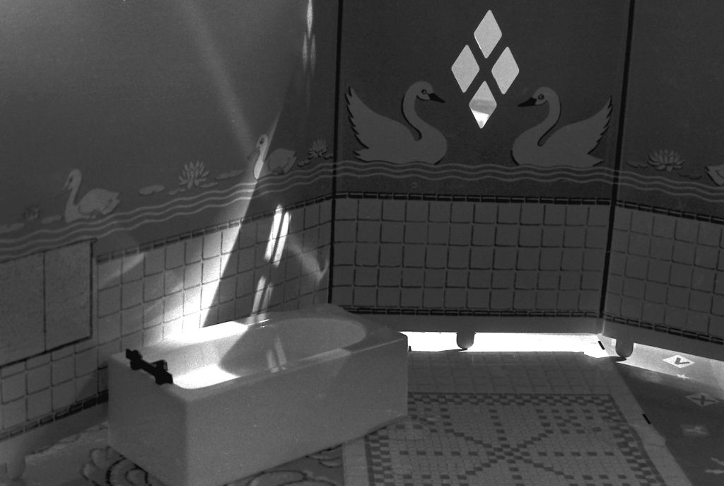 A bathtub stands in a miniature bathroom tiled up to waist height. The upper half of the bathroom is covered with swan-patterned wallpaper. A small amount of light enters the room through four diamond-shaped openings. The photograph is rendered in black and white.