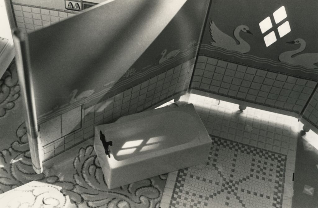 The black-and-white photograph shows the interior of a miniature bathroom. The floor is made of papers imitating different types of flooring. The wall is a standee with printed tiles up to waist height, while the upper section is covered with swan-patterned wallpaper. A miniature bathtub stands in the bathroom.
