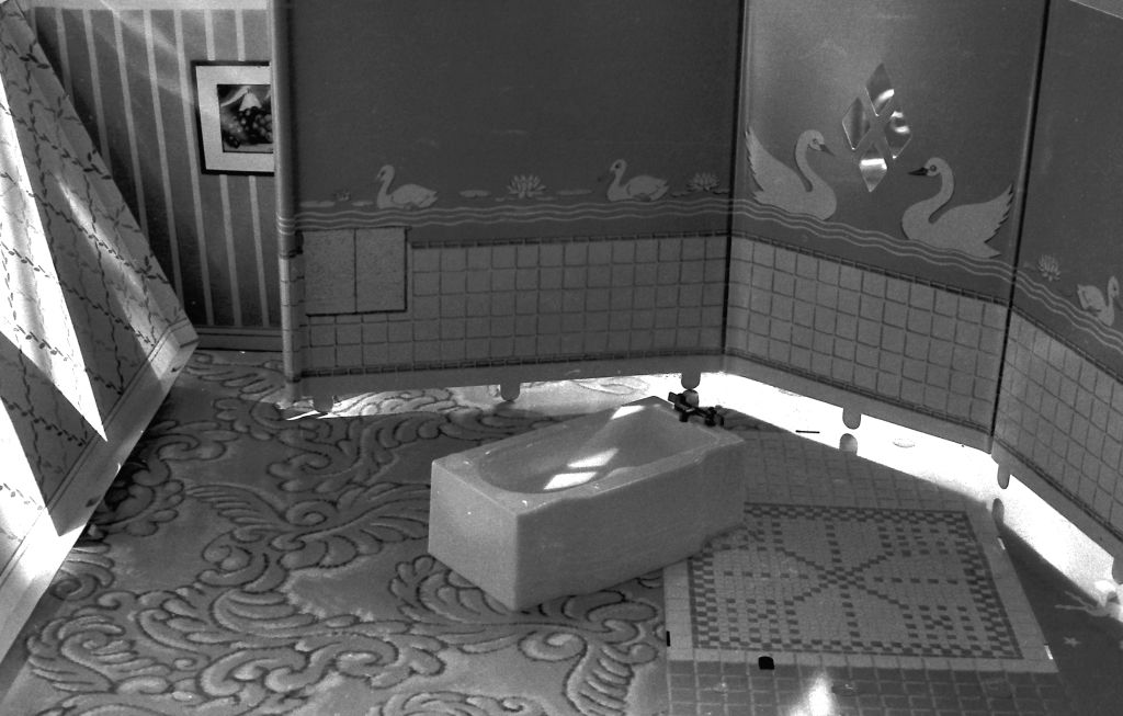 The black-and-white photograph shows the interior of a miniature bathroom. The floor is made of papers imitating different types of flooring. The wall is a standee with printed tiles up to waist height, while the upper section is covered with swan-patterned wallpaper. A miniature bathtub stands at an angle in the bathroom. At the left edge of the image, another standee wall can be seen, tilted and on the verge of falling over.