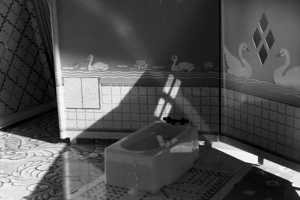 The black-and-white photograph shows the interior of a miniature bathroom. The floor is made of papers imitating different types of flooring. The wall is a standee with printed tiles up to waist height, while the upper section is covered with swan-patterned wallpaper. A miniature bathtub stands at an angle in the bathroom. At the left edge of the image, another standee wall can be seen, tilted and on the verge of falling over. The room appears quite dark, and sparse light creates strong shadows.