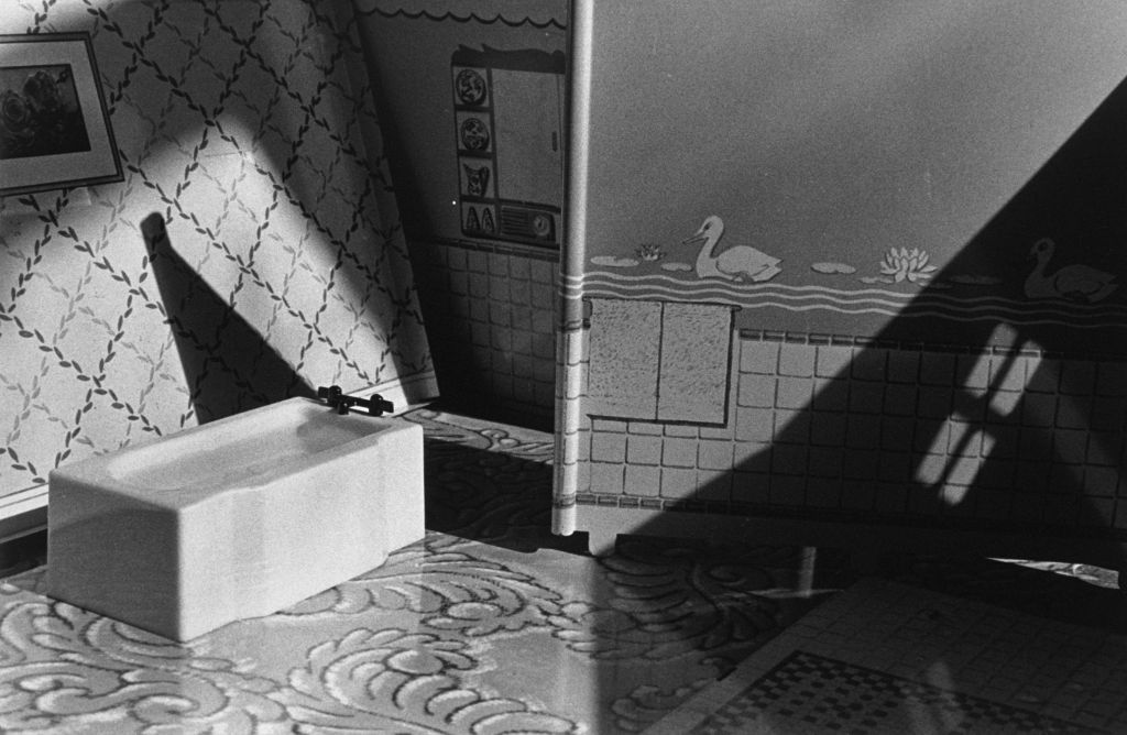 The black-and-white photograph shows the interior of a miniature bathroom. The floor is made of papers imitating different types of flooring. The room is defined by paper partition walls. One wall is tiled and covered above waist height with swan-patterned wallpaper. The other wall features a floral check pattern and stands at an angle within the frame. The bathtub is positioned close to the check-patterned wall, which suggests a living-room context rather than a bathroom setting.