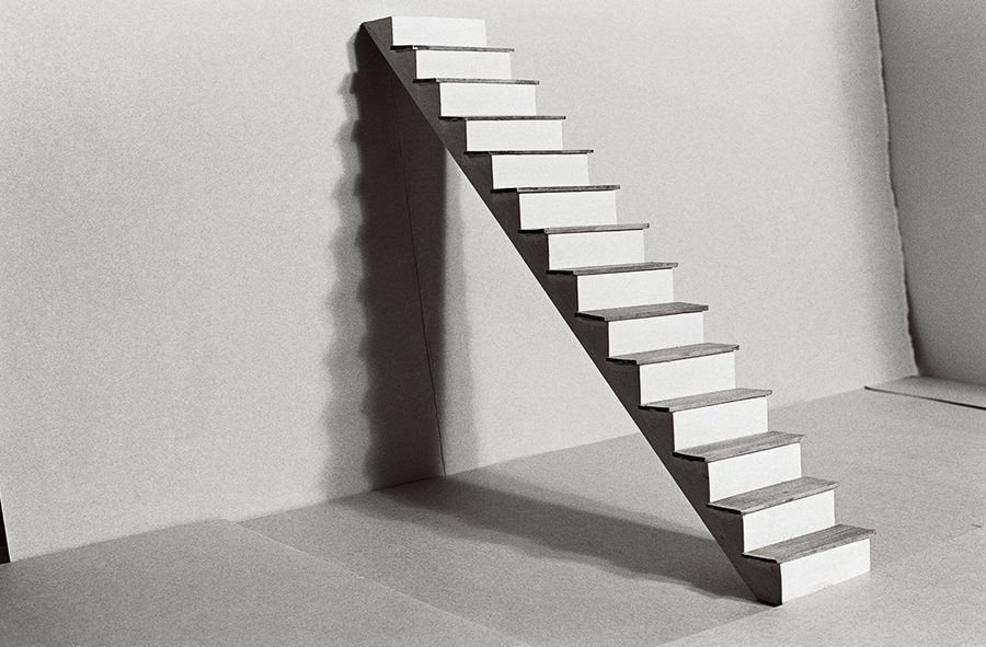 A miniature staircase leans alone within a cardboard room. It casts a distinct shadow. The photograph is rendered in black and white.