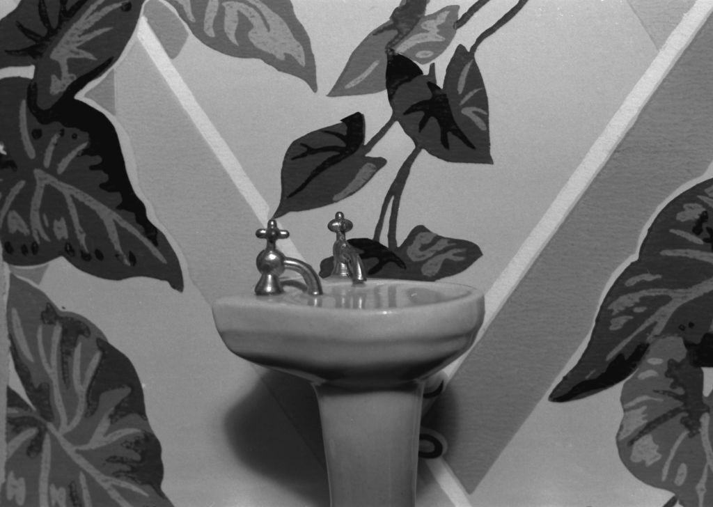 The black-and-white photograph shows a miniature washbasin with two faucets in front of floral wallpaper.