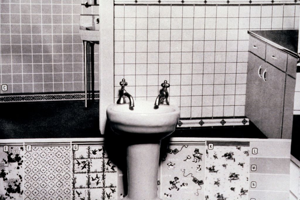 The black-and-white photograph shows a miniature washbasin with two faucets in front of an excerpt from an advertising catalogue. The catalogue features various tile designs as well as sample rooms.