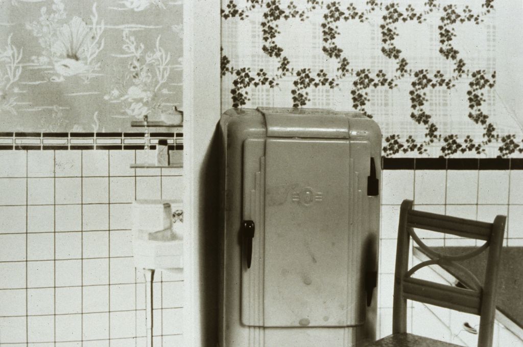 The black-and-white photograph shows a miniature refrigerator and a miniature chair in front of magazine images of interiors. The arrangement creates the impression that the miniature furniture blends seamlessly into the photographs.