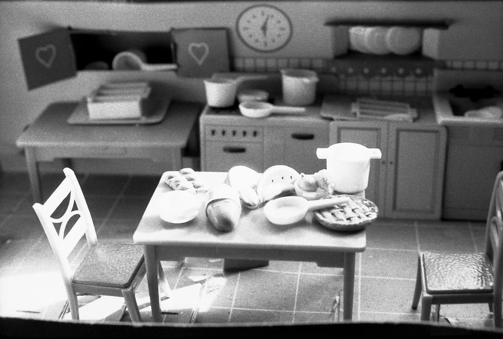 The black-and-white photograph shows the interior of a dollhouse kitchen. In the background, a kitchen unit is visible in soft focus. In the foreground, a table is shown with various foods, including a cake, scattered across it.