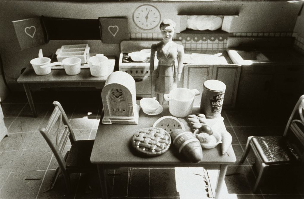 The black-and-white photograph shows the interior of a dollhouse kitchen in which a female doll figure wearing a suit stands. She faces away from a cluttered 1950s-style kitchen unit and looks toward a dining table with scattered food items, including a can of Heinz Baked Beans and a cake.