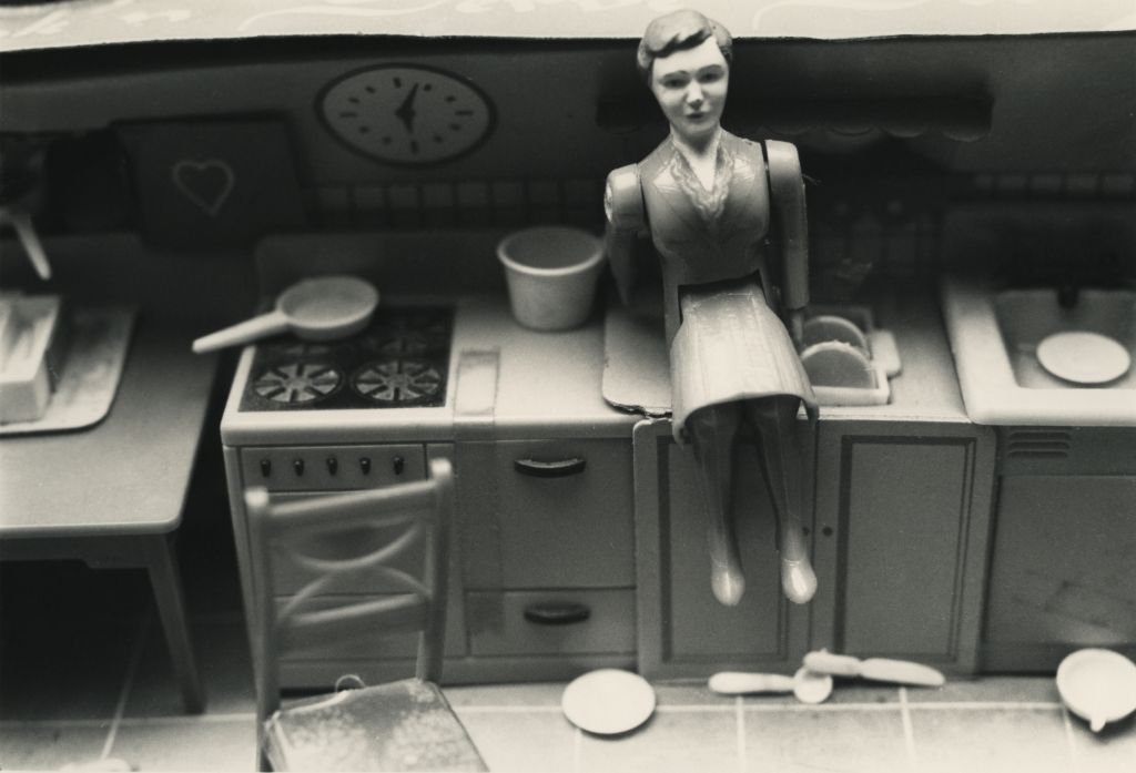 A female doll figure sits in the miniature kitchen of a dollhouse. She is positioned between a set table in the foreground and a cluttered kitchen unit in the background. She sits turned to the side and appears to be looking out of a window through which light falls. The photograph is rendered in black and white.